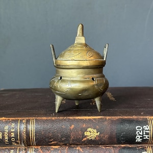 May include: A small, round, brass incense burner featuring three legs, two upright handles, and a removable lid with a pointed finial. The surface of the metal is decorated with etched patterns and small circular cutouts along the upper body.