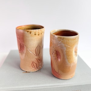 Puede incluir: Dos vasos de cerámica hechos a mano con un diseño de hojas florales. Los vasos son de color melocotón cálido con acentos marrones y naranjas. El diseño está tallado en la superficie. Los vasos miden aproximadamente 10 cm de alto.