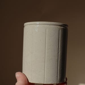 May include: A cylindrical off-white ceramic crock featuring vertical recessed grooves along the exterior body. The vessel has a flat rim and a smooth, matte-textured surface with subtle variations in the clay finish throughout the exterior.