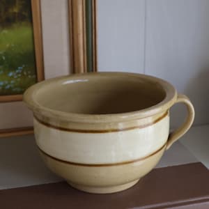 May include: A tan ceramic chamber pot with a single handle and a wide, rounded rim. The exterior features a smooth, glossy finish with two horizontal dark brown stripes encircling the middle section of the bowl, creating a distinct contrast.