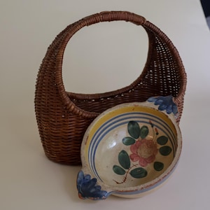 Puede incluir: Una cesta de mimbre trenzado marrón con un asa arqueada alta colocada detrás de un cuenco de cerámica redondo. El cuenco presenta un interior de color crema con una flor rosa pintada a mano, hojas verdes y bordes con rayas azules y dos asas.