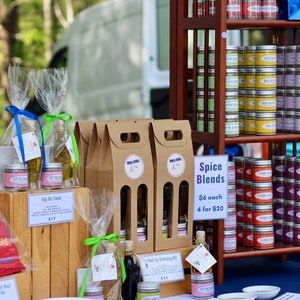 May include: A wooden table display with various spice blends and oil bottles. The spice blends are in small metal tins and are labeled "Spice Blends". The oil bottles are in clear glass bottles with brown paper bags and are labeled "Dip Me Twice". The table is covered in a blue tablecloth. The text "Went Rub You The Right Way" is printed on a red sign.