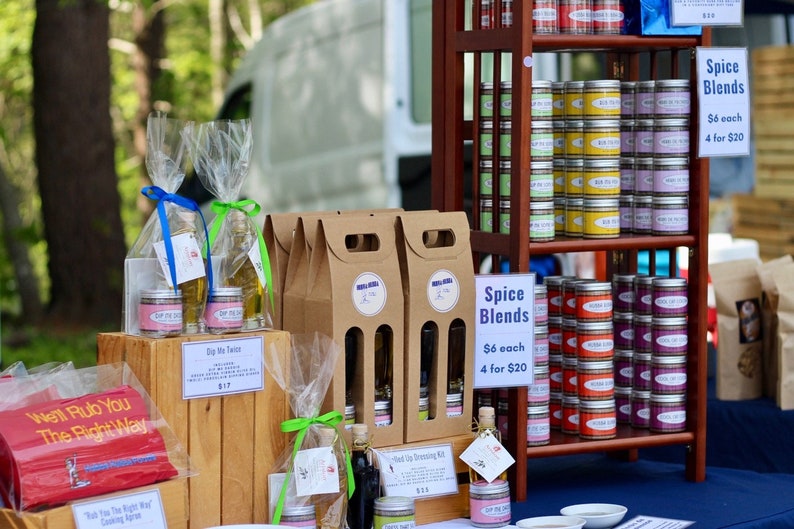 May include: A wooden table display with various spice blends and oil bottles. The spice blends are in small metal tins and are labeled "Spice Blends". The oil bottles are in clear glass bottles with brown paper bags and are labeled "Dip Me Twice". The table is covered in a blue tablecloth. The text "Went Rub You The Right Way" is printed on a red sign.