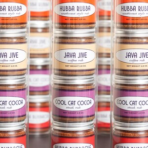 May include: Six clear glass jars with silver lids containing different spice rubs. The jars are stacked in three rows of two. The labels on the jars read "Hubba Rubba Southwest Style Rub", "Java Jive Coffee Rub", "Cool Cat Cocoa Steak Rub", and "Hubba Bubbacue Southern Style Rub".