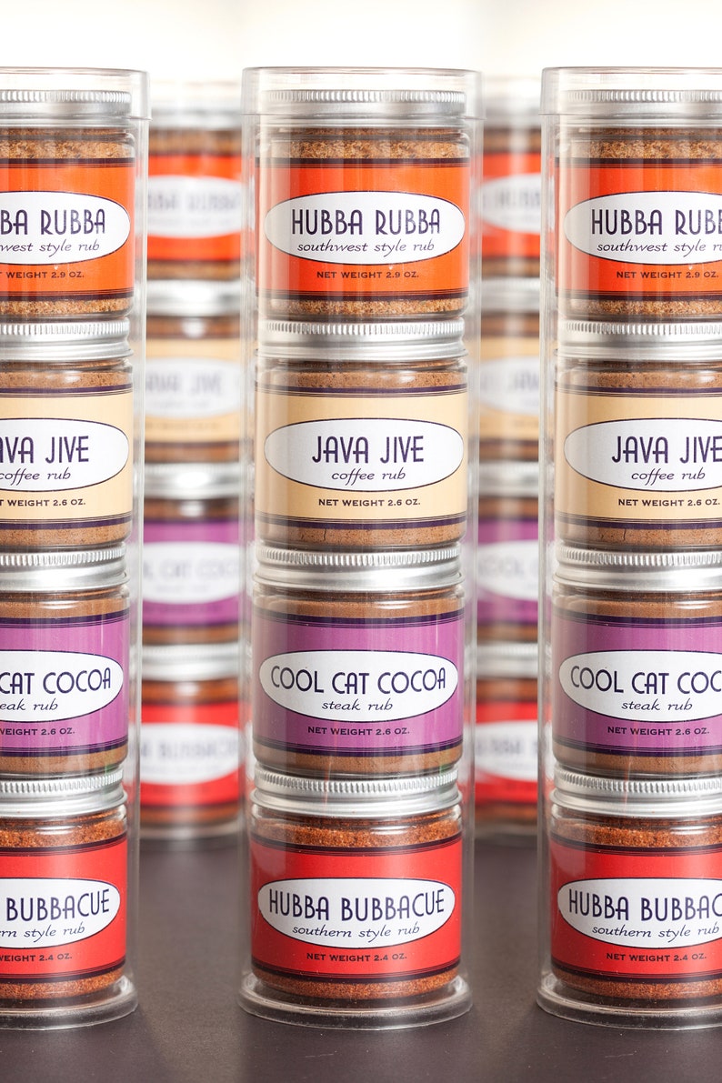 May include: Six clear glass jars with silver lids containing different spice rubs. The jars are stacked in three rows of two. The labels on the jars read "Hubba Rubba Southwest Style Rub", "Java Jive Coffee Rub", "Cool Cat Cocoa Steak Rub", and "Hubba Bubbacue Southern Style Rub".