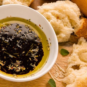 May include: A white bowl filled with a dark brown balsamic vinaigrette dressing with olive oil and herbs. The bowl is on a wooden surface with pieces of crusty bread.