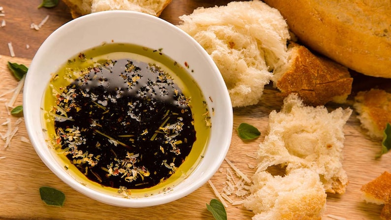 May include: A white bowl filled with a dark brown balsamic vinaigrette dressing with olive oil and herbs. The bowl is on a wooden surface with pieces of crusty bread.