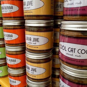 May include: A close-up of a shelf with multiple jars of different flavored steak rubs. The jars are labeled with the names of the rubs, such as "Java Jive Coffee Rub", "Cool Cat Cocoa Steak Rub", and "La Rubba Steak Rub". Each jar has a silver lid and a net weight of 2.6 ounces.