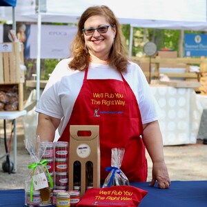 May include: A woman wearing a red apron with the text "We'll Rub You The Right Way" stands behind a table displaying various spice rubs and a gift box. The table is covered in a blue tablecloth.