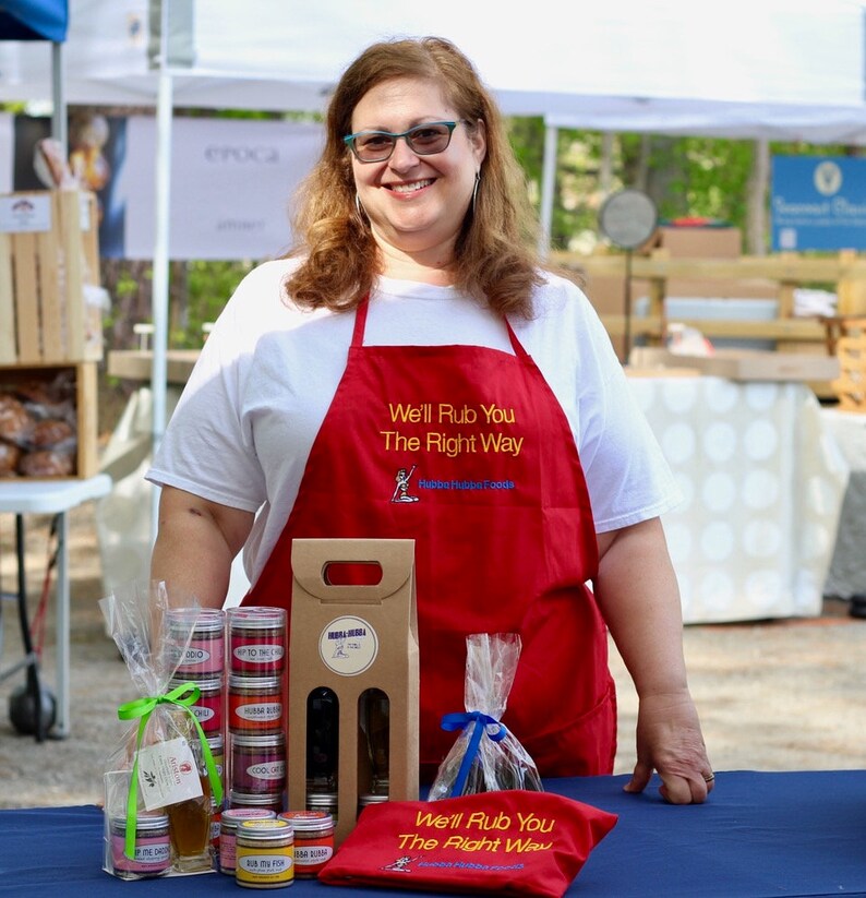 May include: A woman wearing a red apron with the text "We'll Rub You The Right Way" stands behind a table displaying various spice rubs and a gift box. The table is covered in a blue tablecloth.