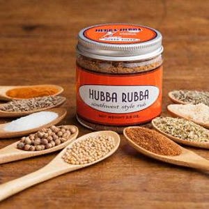 Może przedstawiać: Szklany słoik z przyprawą Hubba Rubba Southwest Style Rub, z srebrną pokrywką. Słoik jest otoczony drewnianymi łyżkami wypełnionymi r&oacute;żnymi przyprawami, w tym solą, nasionami kolendry, nasionami gorczycy i płatkami czerwonej papryki.