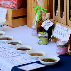 May include: A table display of various salad dressings and oils in small white bowls with labels. The labels include names like "Cool Oil" and "Fish My Friend". There are also bottles of olive oil and balsamic vinegar, along with a small jar of salad seasoning. The text "Dolled Up Dressing Kit" and "$2.25" are visible on a brown paper sign.