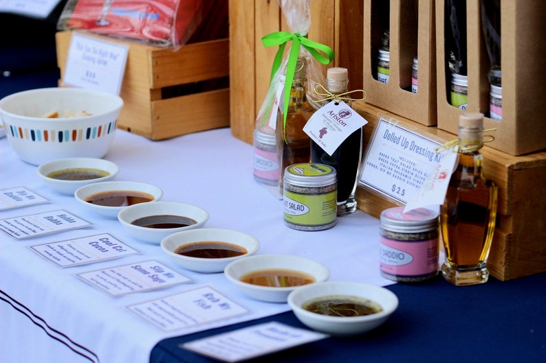 May include: A table display of various salad dressings and oils in small white bowls with labels. The labels include names like "Cool Oil" and "Fish My Friend". There are also bottles of olive oil and balsamic vinegar, along with a small jar of salad seasoning. The text "Dolled Up Dressing Kit" and "$2.25" are visible on a brown paper sign.