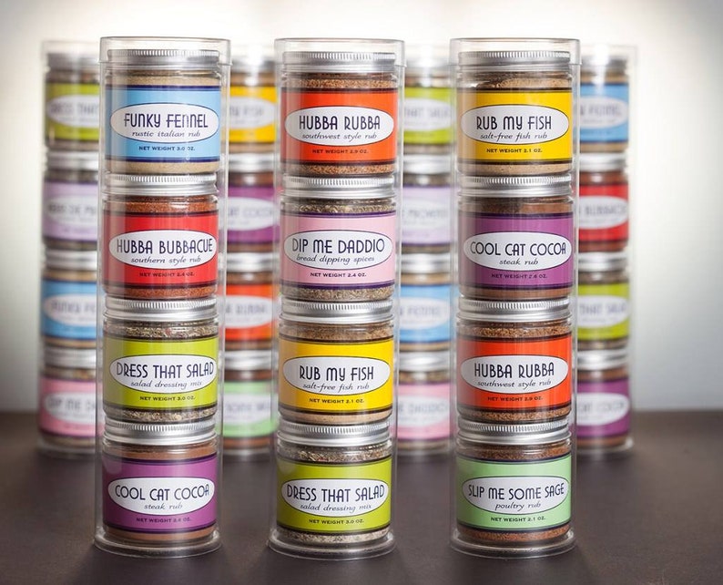 May include: A set of six clear glass jars filled with different spice blends. Each jar has a colorful label with a unique name and description. The names include "Funky Fennel", "Hubba Rubba", "Rub My Fish", "Hubba Bubbacue", "Dip Me Daddo", "Cool Cat Cocoa", "Dress That Salad", "Slip Me Some Sage".