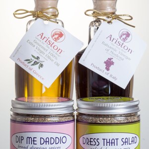 May include: Two glass bottles of olive oil and balsamic vinegar with labels that read "Ariston" and "Gourmet 100% Extra Virgin Olive Oil" and "Balsamic Vinegar of Modena". Two glass jars of spices with labels that read "Dip Me Daddi Bread Dipping Spices" and "Dress That Salad Salad Dressing Mix".
