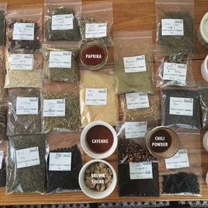 May include: A collection of spices and ingredients in clear plastic bags and small bowls. The ingredients include paprika, cayenne pepper, chili powder, brown sugar, table salt, Himalayan sea salt, cocoa nibs, and coffee beans. The labels on the bags indicate the names of the spices and ingredients.