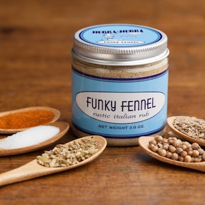 May include: A glass jar with a silver lid filled with a light brown spice blend. The label reads "Funky Fennel Rustic Italian Rub" and "Net Weight 3.0 oz". The jar is surrounded by small wooden spoons filled with various spices, including red pepper flakes, salt, fennel seeds, and coriander seeds.