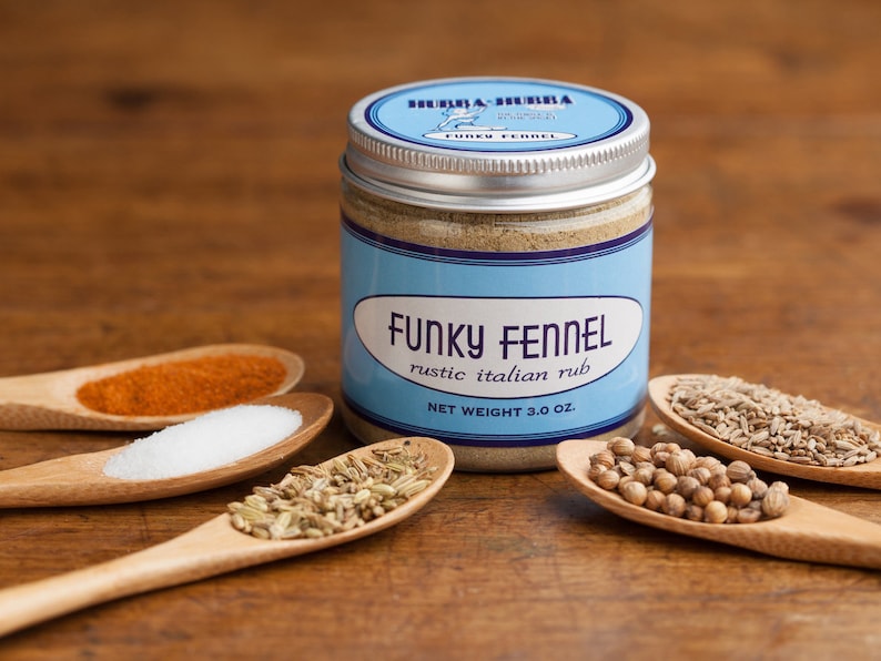 May include: A glass jar with a silver lid filled with a light brown spice blend. The label reads "Funky Fennel Rustic Italian Rub" and "Net Weight 3.0 oz". The jar is surrounded by small wooden spoons filled with various spices, including red pepper flakes, salt, fennel seeds, and coriander seeds.