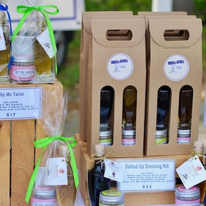 May include: A wooden crate with two glass bottles of olive oil and two small jars of dip mix. The crate is labeled "Dip Me Twice" and the price is $17.  Next to the crate are three brown cardboard boxes with two glass bottles and two small jars in each box. The boxes are labeled "Dolled Up Dressing Kit" and the price is $25.  A sign on the right side of the image reads "Spice Blends" with the prices $6 each and 4 for $20.