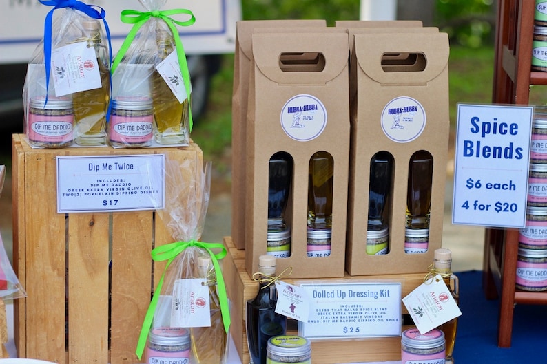 May include: A wooden crate with two glass bottles of olive oil and two small jars of dip mix. The crate is labeled "Dip Me Twice" and the price is $17.  Next to the crate are three brown cardboard boxes with two glass bottles and two small jars in each box. The boxes are labeled "Dolled Up Dressing Kit" and the price is $25.  A sign on the right side of the image reads "Spice Blends" with the prices $6 each and 4 for $20.