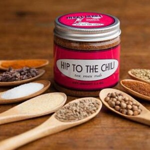 Może przedstawiać: Szklany słoik ze srebrną pokrywką wypełniony brązową mieszanką przypraw. Etykieta zawiera napis "Hip to the Chili" i "Tex Mex Rub". Słoik jest otoczony małymi drewnianymi łyżeczkami zawierającymi r&oacute;żne przyprawy, w tym kminek, kolendra, s&oacute;l, czosnek w proszku, papryka chili i oregano.