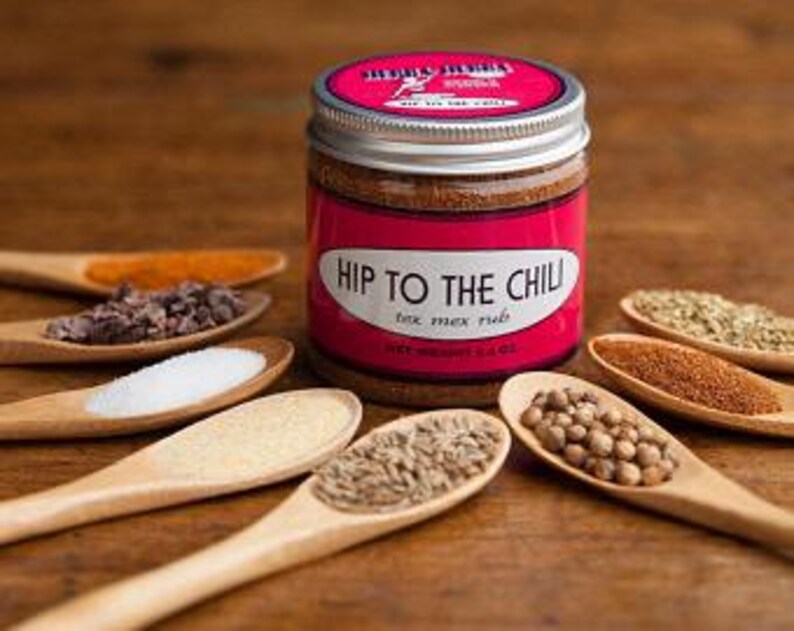 Może przedstawiać: Szklany słoik ze srebrną pokrywką wypełniony brązową mieszanką przypraw. Etykieta zawiera napis "Hip to the Chili" i "Tex Mex Rub". Słoik jest otoczony małymi drewnianymi łyżeczkami zawierającymi r&oacute;żne przyprawy, w tym kminek, kolendra, s&oacute;l, czosnek w proszku, papryka chili i oregano.