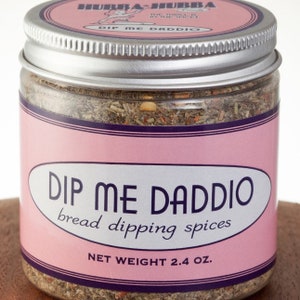 May include: A glass jar with a silver lid filled with brown and red spices. The label is pink with black text that reads "Dip Me Daddio bread dipping spices" and "Net Weight 2.4 oz."