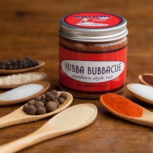 Może przedstawiać: Szklany słoik z przyprawą Hubba Bubbacue Southern Style Rub, z czerwoną etykietą i białym tekstem. Słoik otoczony jest drewnianymi łyżkami wypełnionymi r&oacute;żnymi przyprawami, w tym czarnymi ziarnami pieprzu, solą i papryką.
