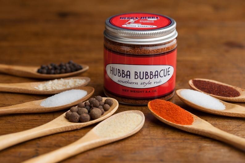 Może przedstawiać: Szklany słoik z przyprawą Hubba Bubbacue Southern Style Rub, z czerwoną etykietą i białym tekstem. Słoik otoczony jest drewnianymi łyżkami wypełnionymi r&oacute;żnymi przyprawami, w tym czarnymi ziarnami pieprzu, solą i papryką.