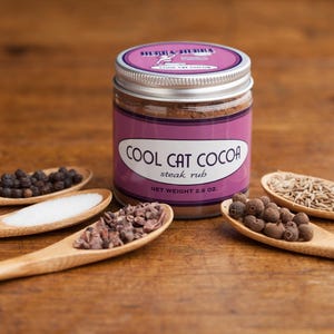 Może przedstawiać: Szklany słoik ze srebrną pokrywką wypełniony brązową mieszanką przypraw. Etykieta zawiera napis "Cool Cat Cocoa Steak Rub" i "Waga netto 2,8 oz". Słoik otoczony jest drewnianymi łyżkami wypełnionymi r&oacute;żnymi przyprawami, w tym czarnymi ziarnami pieprzu, solą, nibsami kakaowymi, jagodami pimento i nasionami kminku.