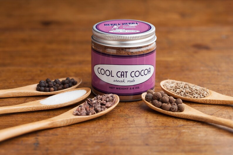 Może przedstawiać: Szklany słoik ze srebrną pokrywką wypełniony brązową mieszanką przypraw. Etykieta zawiera napis "Cool Cat Cocoa Steak Rub" i "Waga netto 2,8 oz". Słoik otoczony jest drewnianymi łyżkami wypełnionymi r&oacute;żnymi przyprawami, w tym czarnymi ziarnami pieprzu, solą, nibsami kakaowymi, jagodami pimento i nasionami kminku.