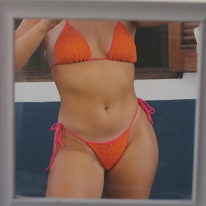 Könnte beinhalten: Orangener Häkel-Bikini mit rosa Einfassung. Das handgefertigte Triangel-Oberteil und der String-Bikini-Slip sind zu sehen. Die Badebekleidung spiegelt sich in einem Spiegel wider und zeigt die leuchtende Farbe und die detaillierte Naht.