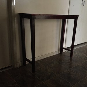 May include: A dark brown wooden console table with a rectangular top and four legs. The table is standing on a dark tiled floor.