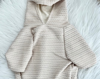 May include: A cream-colored knitted hoodie for a pet. The hoodie has a cable knit pattern and a hood with a drawstring.
