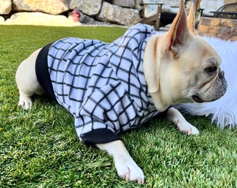 Puede incluir: Un bulldog francés con un suéter de vellón a cuadros blanco y negro con ribete negro. El perro está acostado en césped artificial verde.