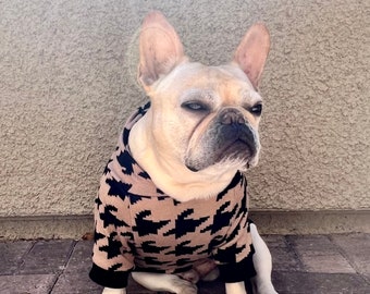 May include: A French bulldog wearing a brown and black houndstooth sweater sits on a brick patio. The dog has a serious expression on its face.