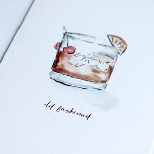 May include: Watercolour illustration of an Old Fashioned cocktail in a rocks glass with ice, cherries, and an orange slice. The words "old fashioned" are written in a burgundy script below the glass.