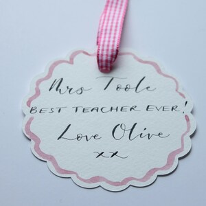 May include: A white card with pink watercolour edges and a handwritten message that says "Mrs. Toole Best Teacher Ever! Love Olive xx".