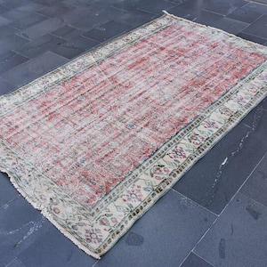 May include: Rectangular vintage rug with a faded red and cream color palette. The rug features a central field of red with a distressed appearance, framed by a cream border with floral and geometric patterns. The rug is placed on a dark gray tiled floor.