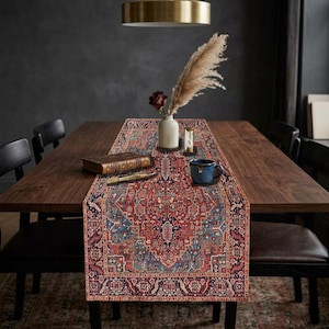 May include: A patterned table runner in red, blue, and cream, on a dark wood dining table. The runner has a detailed, traditional design. A vase with dried flowers, a book, and a blue mug are on the table. The table is surrounded by black chairs.