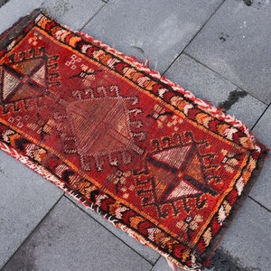 May include: A red, orange, and black patterned rug with a fringed edge. The rug has a geometric design with a central diamond shape.