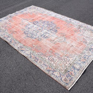 May include: Rectangular vintage rug with a distressed appearance. The rug has a central field of faded coral, framed by a border of blue and cream floral patterns. It is placed on a dark asphalt surface.