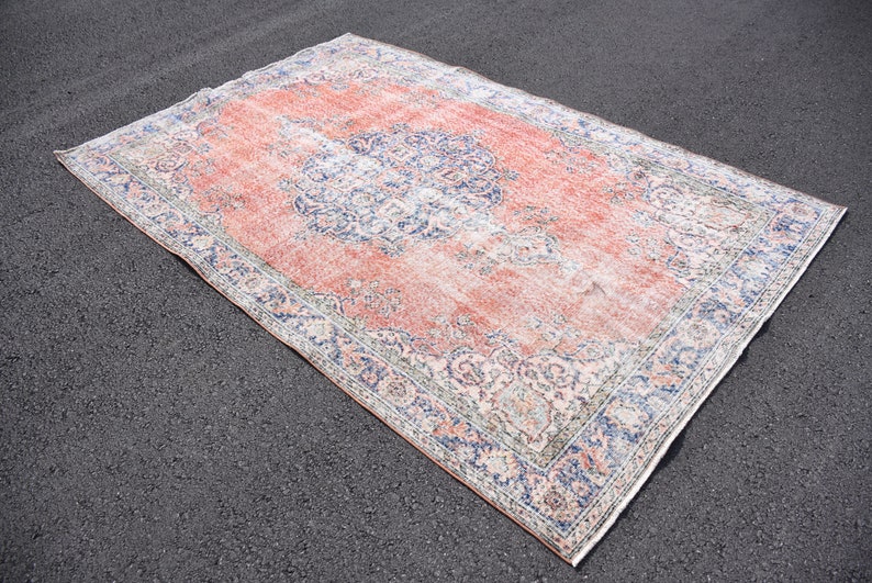 May include: Rectangular vintage rug with a distressed appearance. The rug has a central field of faded coral, framed by a border of blue and cream floral patterns. It is placed on a dark asphalt surface.
