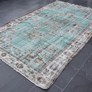 May include: A rectangular, distressed-look rug with a central turquoise field and a detailed floral pattern. The rug has a cream-colored border with brown and beige accents, creating a vintage aesthetic. The rug is placed on a dark gray tiled floor.