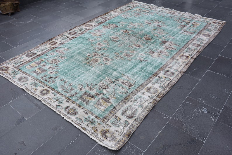 May include: A rectangular, distressed-look rug with a central turquoise field and a detailed floral pattern. The rug has a cream-colored border with brown and beige accents, creating a vintage aesthetic. The rug is placed on a dark gray tiled floor.