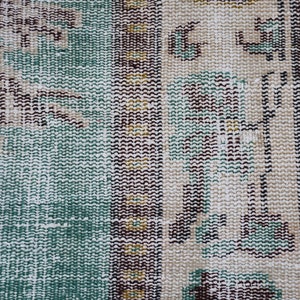 May include: Close-up of a section of a vintage rug with a distressed, textured appearance. The rug features a color palette of teal, beige, brown, and touches of yellow. The design includes geometric and floral patterns.