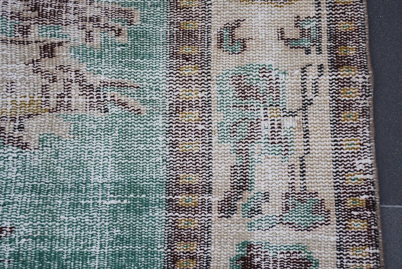 May include: Close-up of a section of a vintage rug with a distressed, textured appearance. The rug features a color palette of teal, beige, brown, and touches of yellow. The design includes geometric and floral patterns.