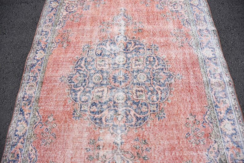 May include: A distressed, rectangular rug with a central medallion design. The rug has a faded, vintage aesthetic with a coral-red field and a navy blue and cream border and medallion. The rug's dimensions are not visible.
