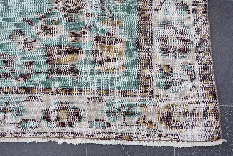 May include: Close-up of a distressed, vintage rug with a faded, floral pattern. The rug features a teal, beige, and brown color palette. The design includes floral and geometric elements, with a fringed edge.