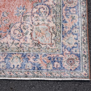 May include: A fragment of a vintage rug with a faded, distressed look. The rug displays a central field of muted coral with detailed floral patterns in blue, cream, and green. A detailed border frames the design.